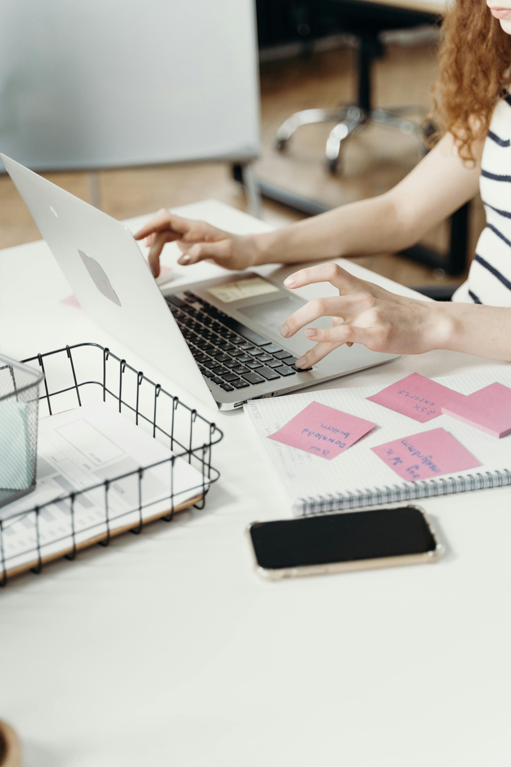 A woman working on a laptop, surrounded by sticky notes, paper, and a phone in a modern office setting.